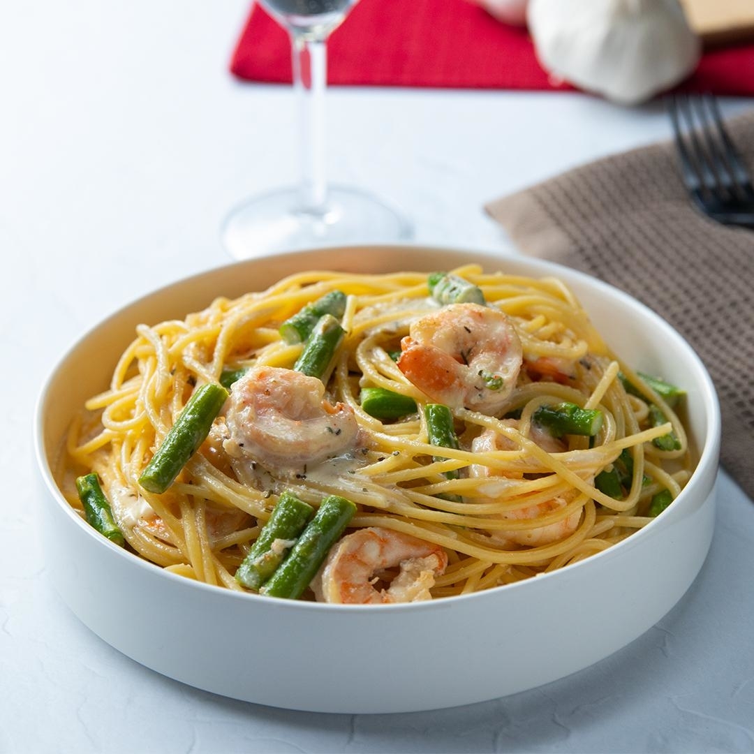A bowl of pasta with shrimp and green beans in a creamy sauce, placed on a table with a wine glass, garlic bulb, fork, and napkin in the background