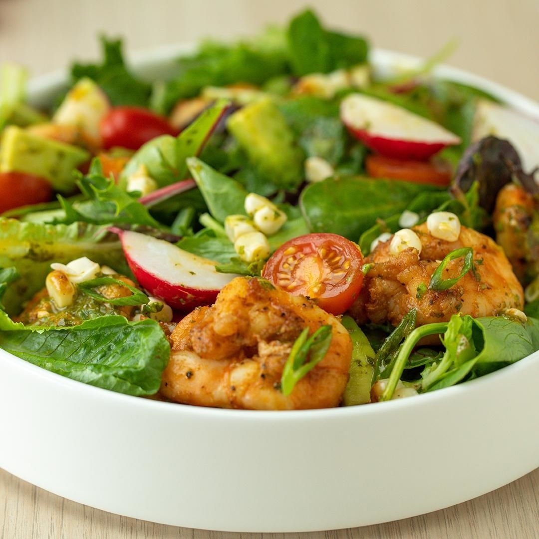 A fresh salad with mixed greens, cherry tomatoes, radishes, grilled shrimp, avocado, corn, and herbs in a white bowl