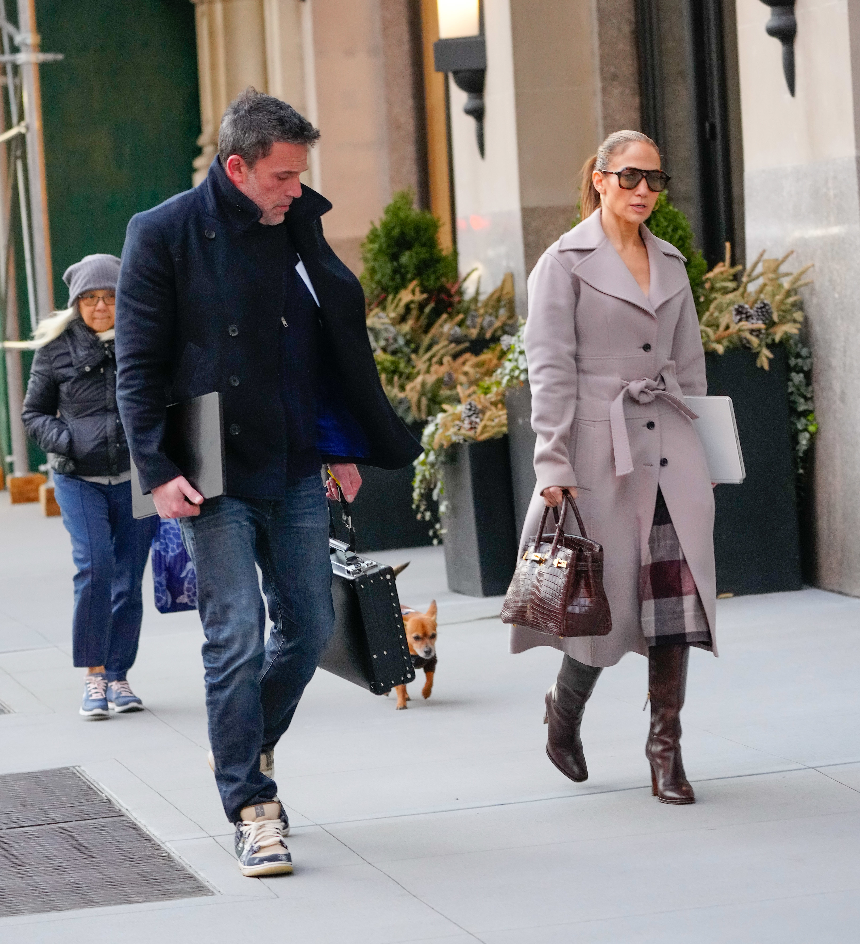 Ben Affleck and Jennifer Lopez walk on a city sidewalk, wearing stylish outerwear. They are accompanied by an unidentified person and a small dog
