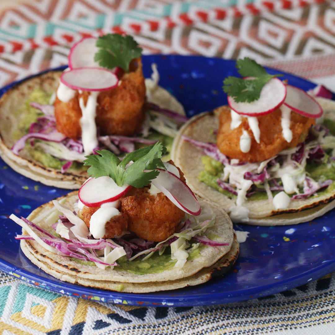 Three fish tacos topped with sliced radishes, cilantro, shredded cabbage, and drizzled with white sauce on a blue plate