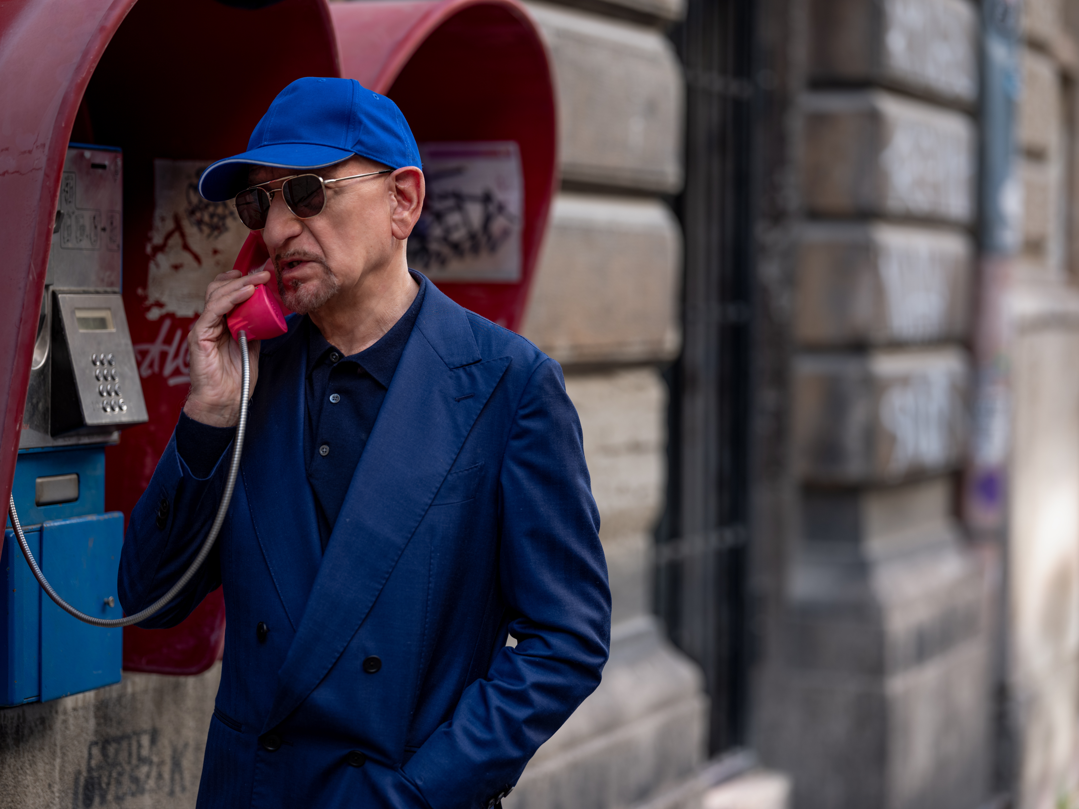 Ben Kingsley in a blue suit and cap, using a red public phone on a city street