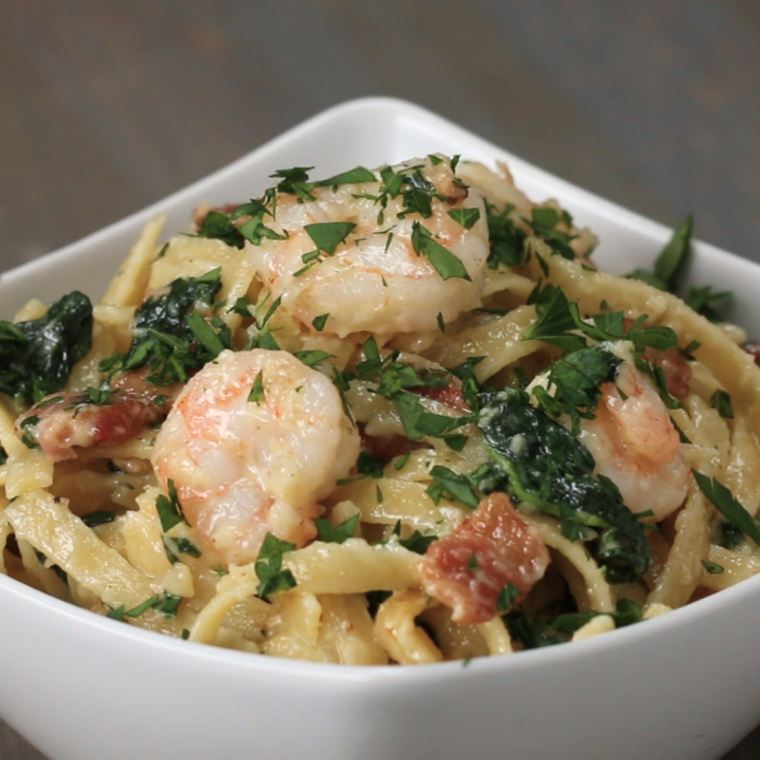 A close-up of a bowl of pasta with shrimp, spinach, bacon, and parsley garnish