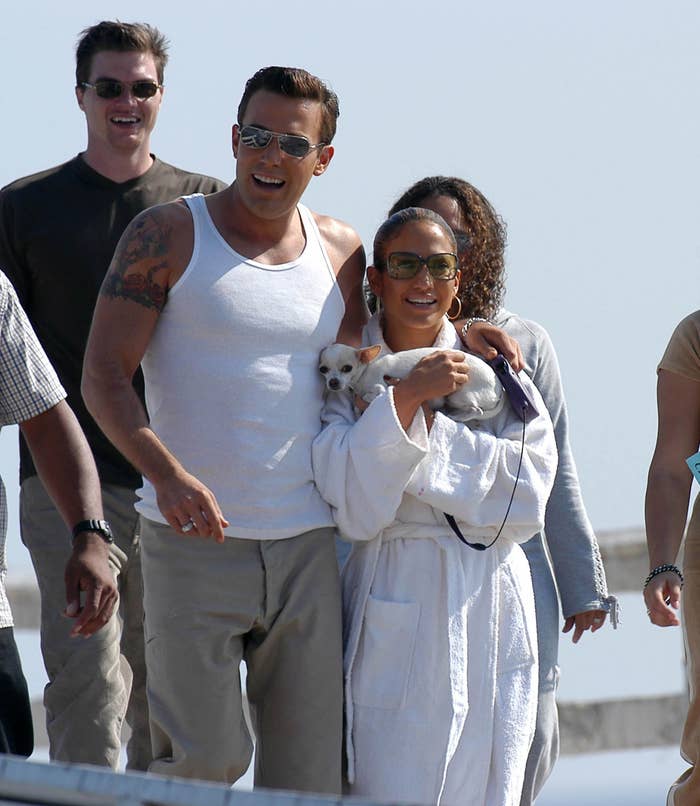 Ben Affleck and Jennifer Lopez walking outside, smiling and wearing casual clothes. Jennifer holds a small dog. Several people are seen in the background