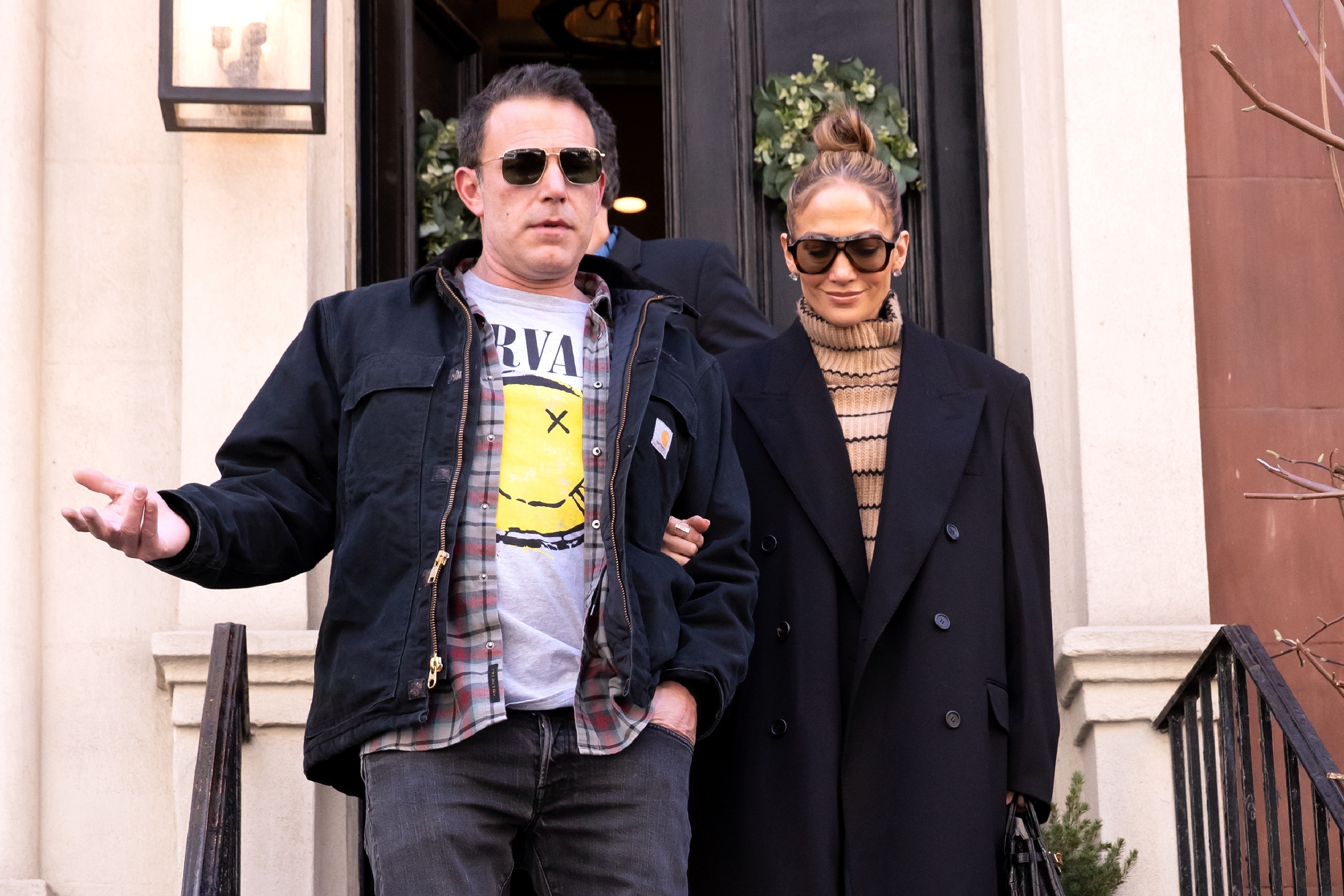Ben Affleck and Jennifer Lopez leaving a building, both wearing sunglasses and casual outfits; Ben Affleck in a Nirvana shirt and jacket, Jennifer Lopez in a black coat