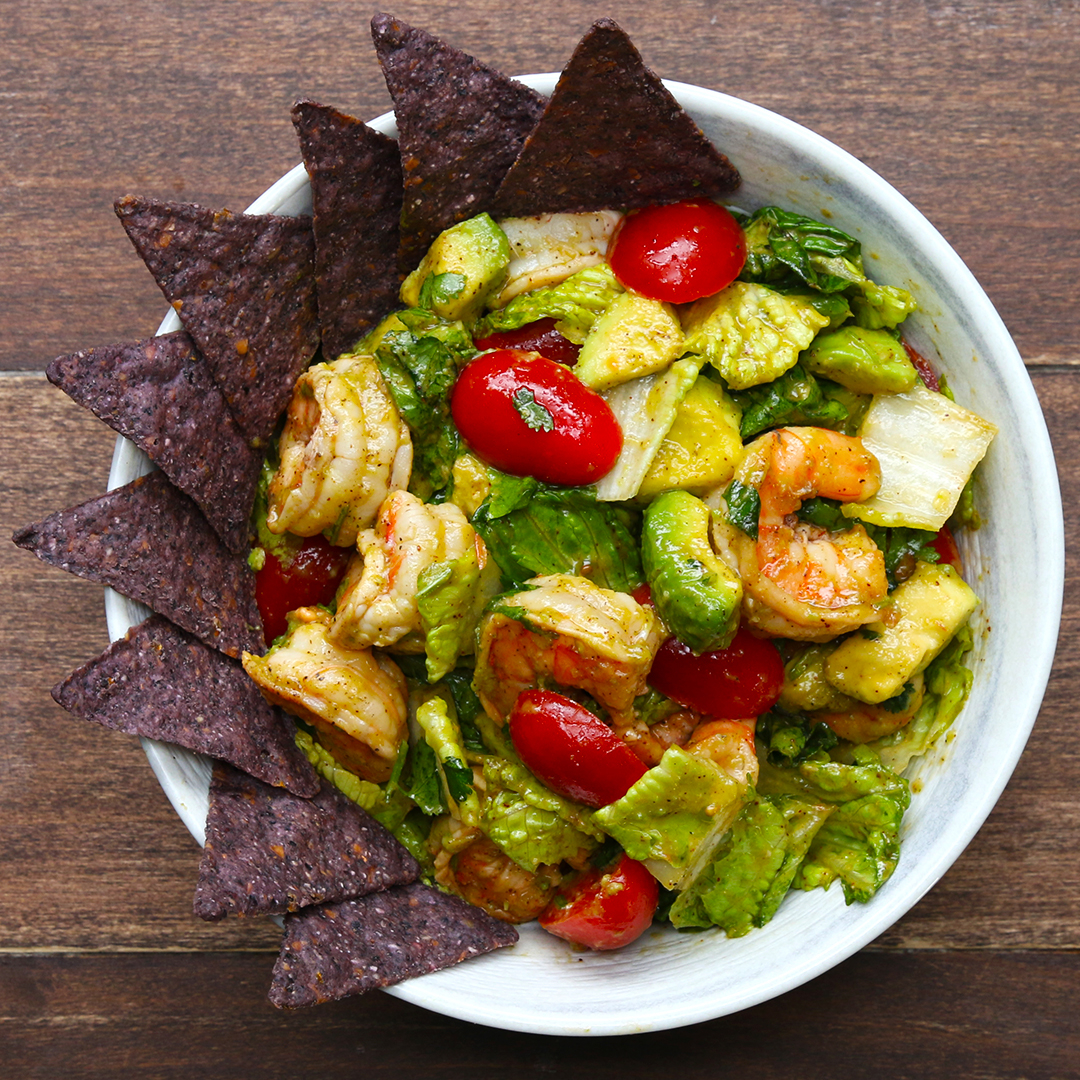 A bowl of shrimp avocado salad garnished with cherry tomatoes, lettuce, and blue corn tortilla chips on the side