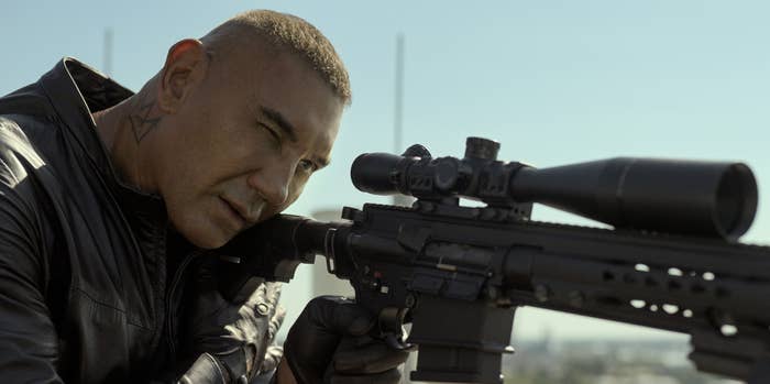 Dave Bautista aims a sniper rifle while wearing a black leather jacket and gloves