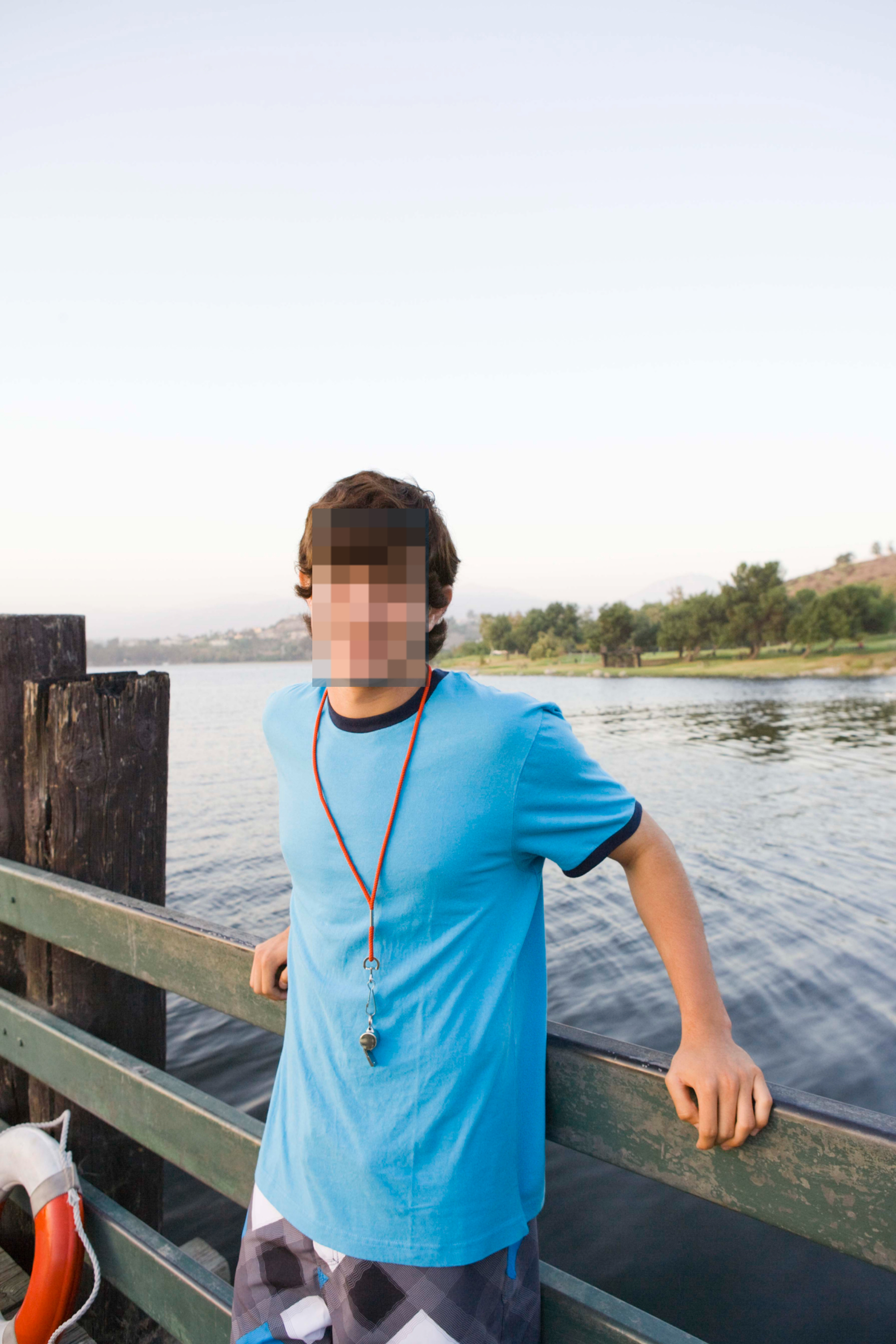 A young man, smiling, stands by a lakeside fence wearing a shirt with a whistle necklace. Trees and water are in the background