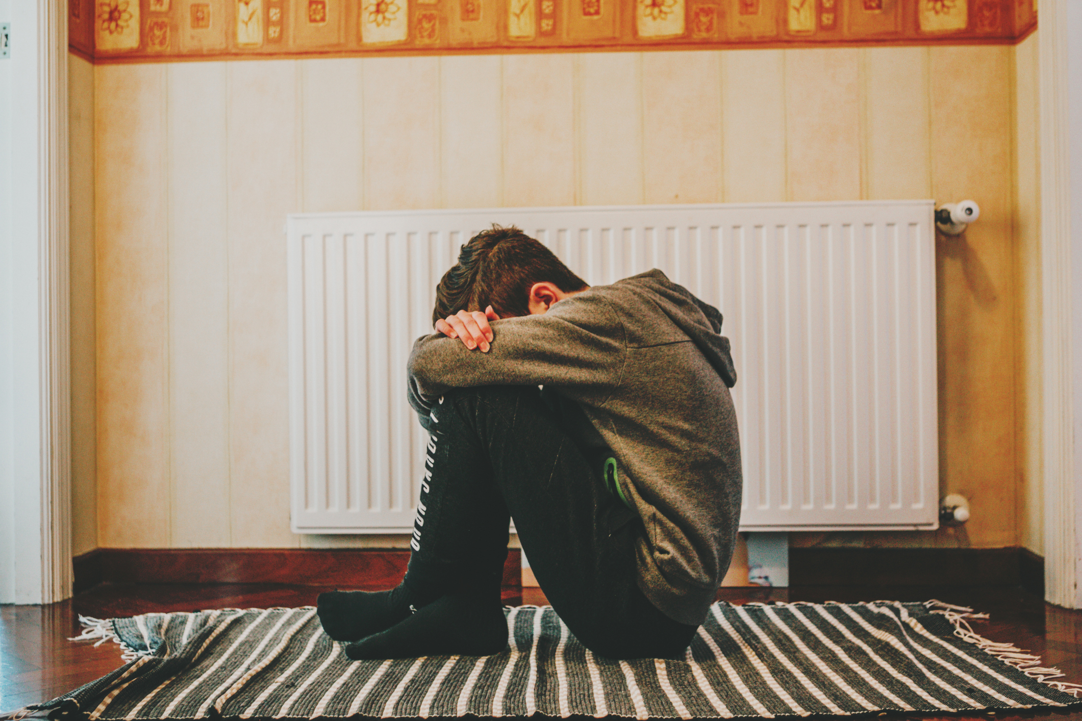A person sits alone on a striped rug, hugging their knees, positioned in front of a radiator. Their face is not visible