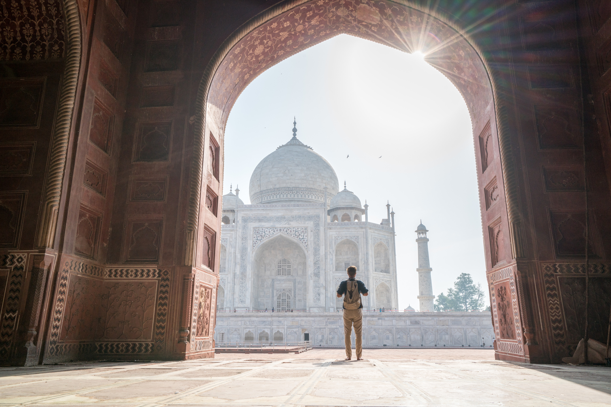 Person stands under an archway, gazing at the Taj Mahal in Agra, India, with the sun shining above the iconic monument