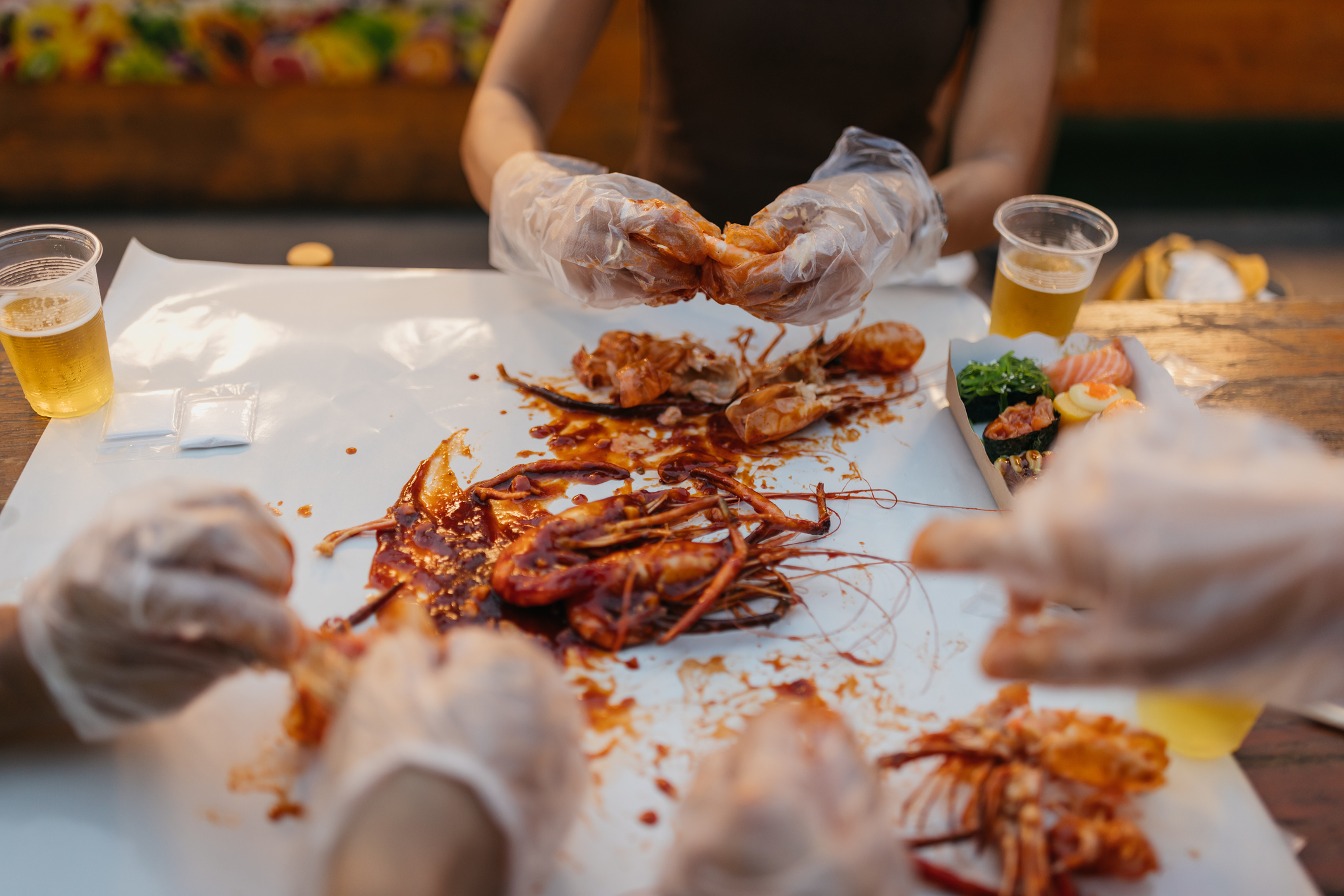 People enjoying a seafood meal with gloves, cracking shells, and sipping on drinks outdoors. The image focuses on hands, food, and drinks