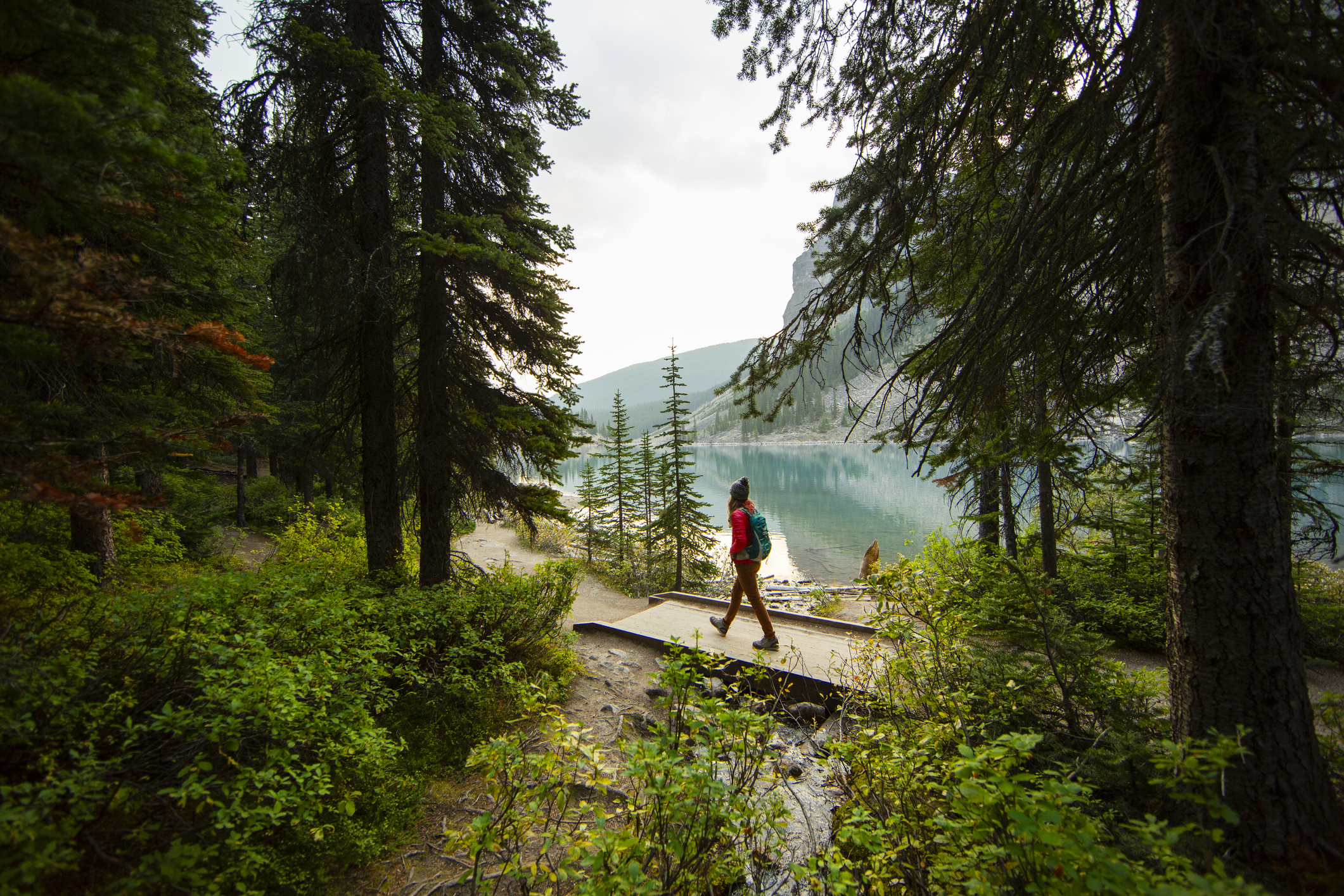 A hiker walks on a forest trail near a serene lake surrounded by trees and mountains