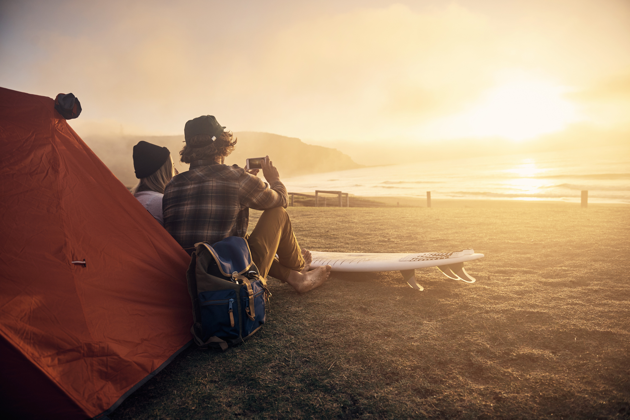 Two people are sitting by a tent on a coastal hill, admiring the sunset with a surfboard nearby, suggesting a camping trip and surfing adventure