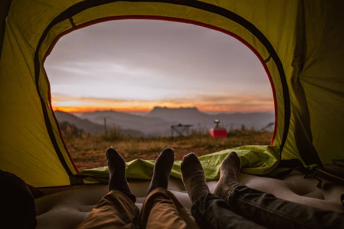 Two people inside a tent with their feet sticking out, overlooking a scenic mountain view at dawn. Camping gear and misty mountains visible in the background