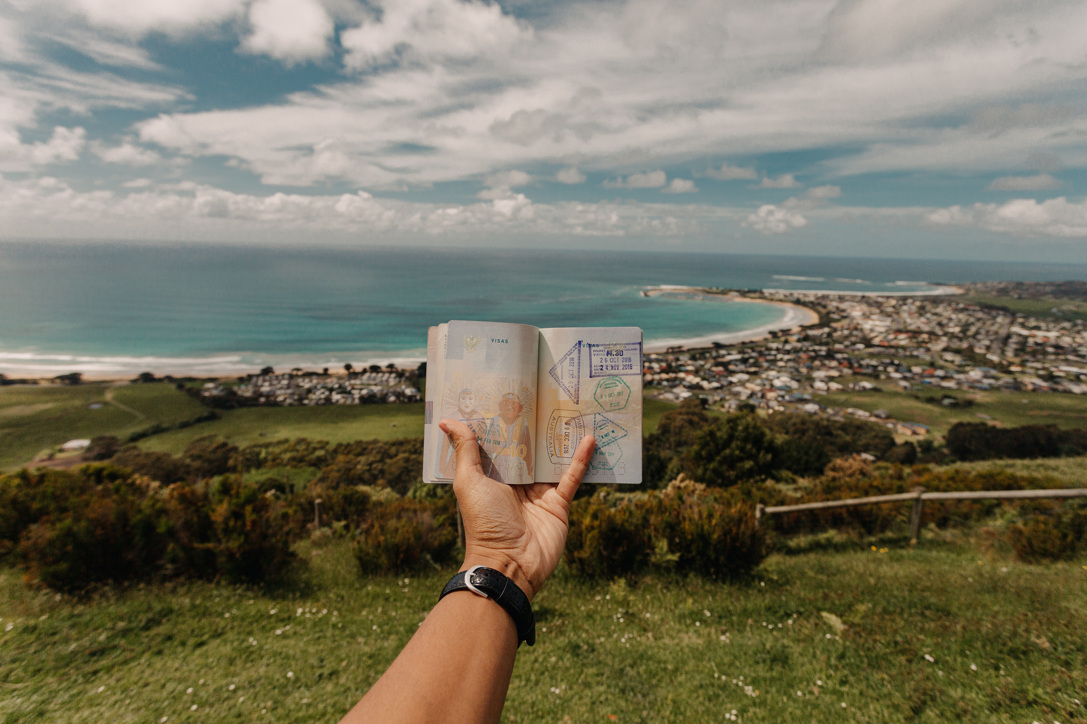 Person holding an open passport with visa stamps against a scenic coastal landscape, featuring a shoreline, ocean, and a town