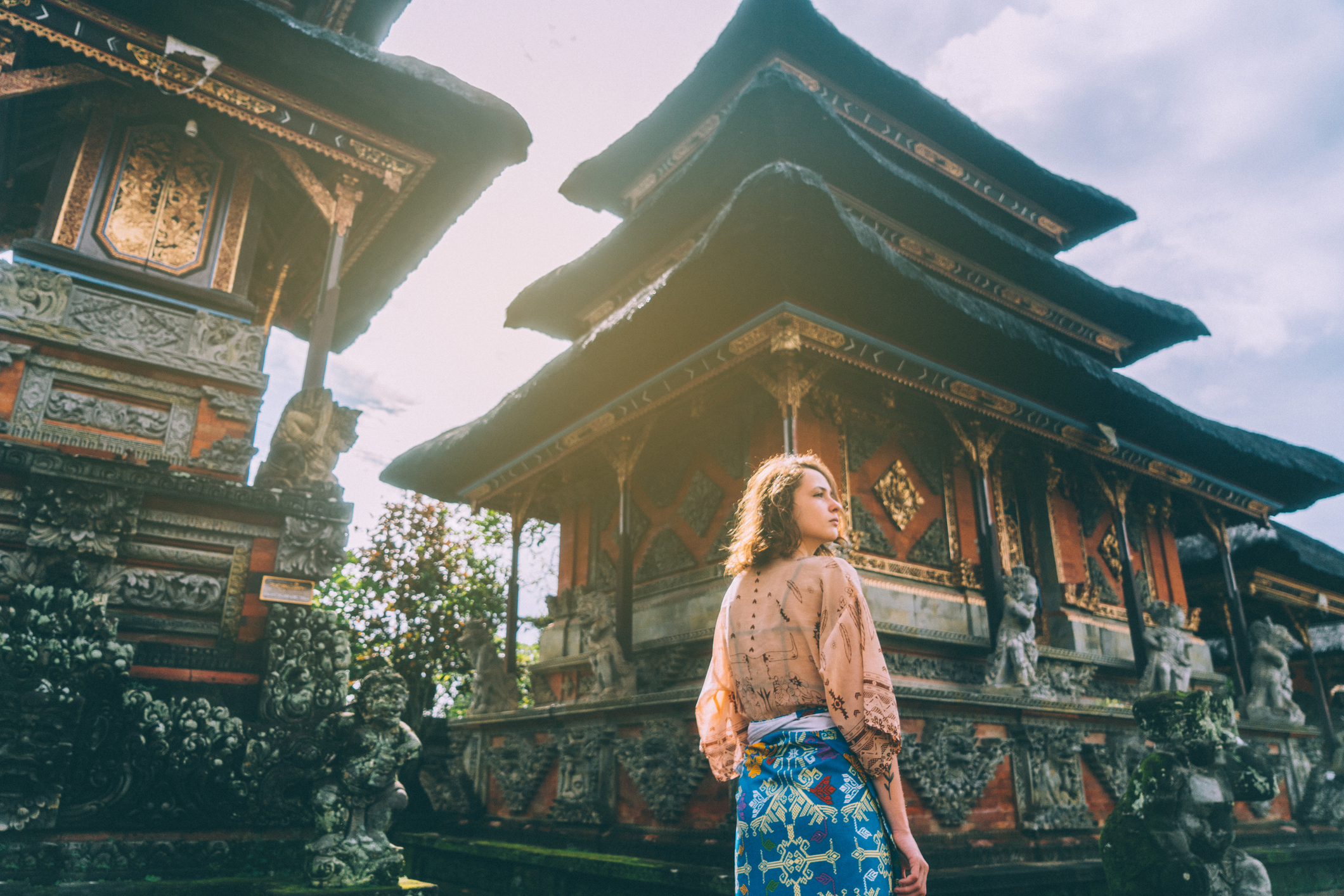 A woman in a patterned skirt and sheer blouse stands before ornate traditional Balinese temples, gazing upwards. The background features intricate temple details