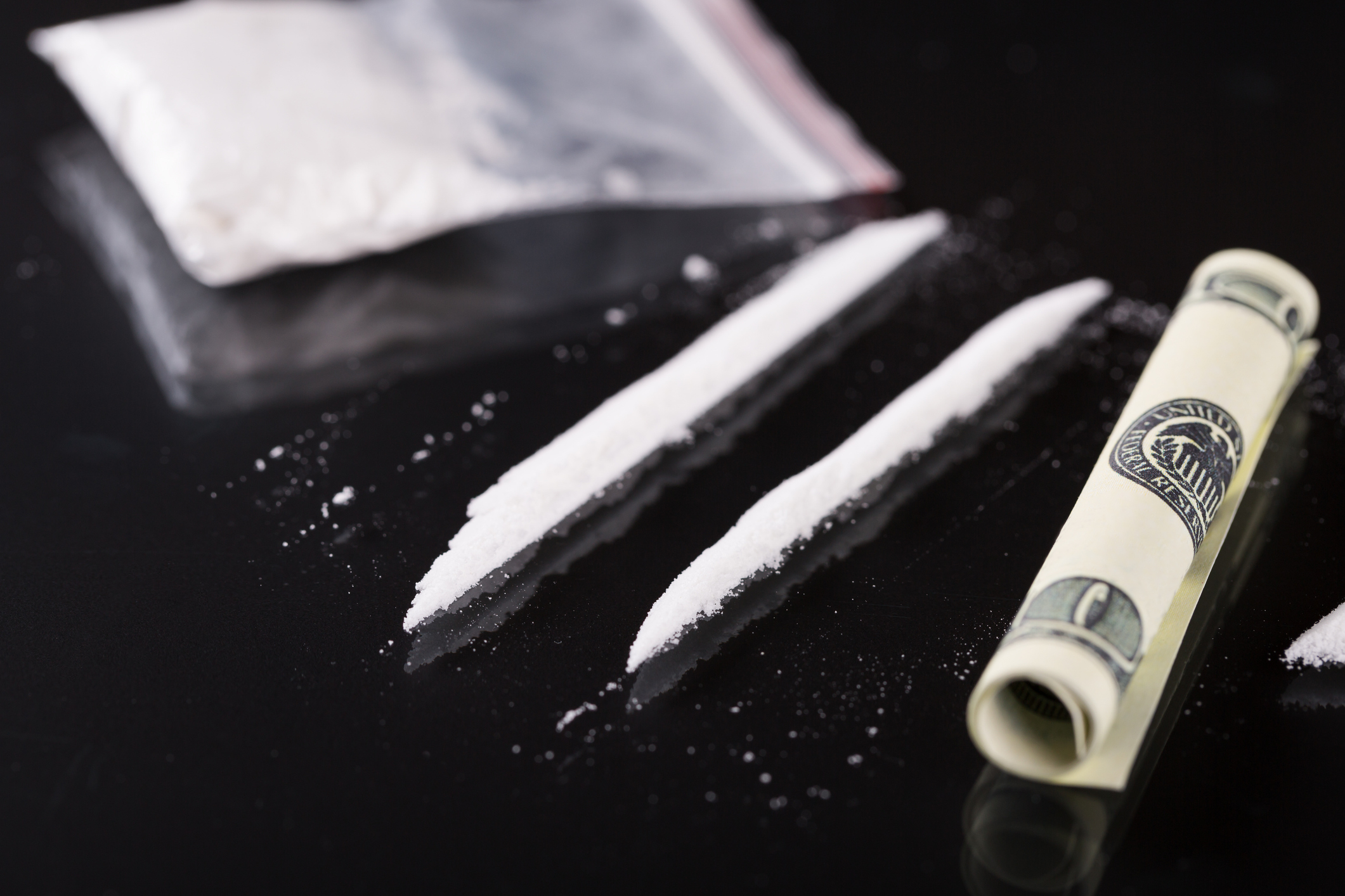 Image of two lines of white powder and a rolled-up dollar bill on a black surface, with a small plastic bag containing more powder at the top