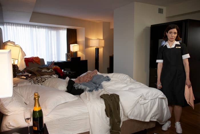 Hotel maid stands beside an unmade bed in a cluttered room, with clothes and items strewn about. A bottle of champagne and a glass are seen on the bedside table