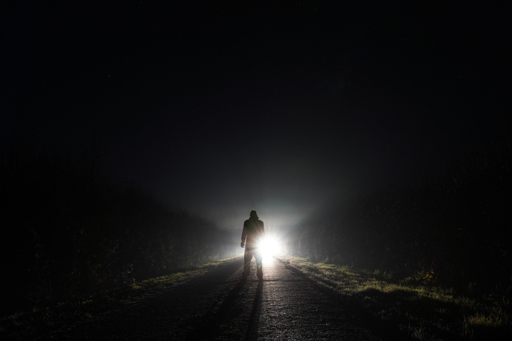 A person stands alone on a dark road facing an oncoming bright light in the distance, creating a dramatic silhouette effect