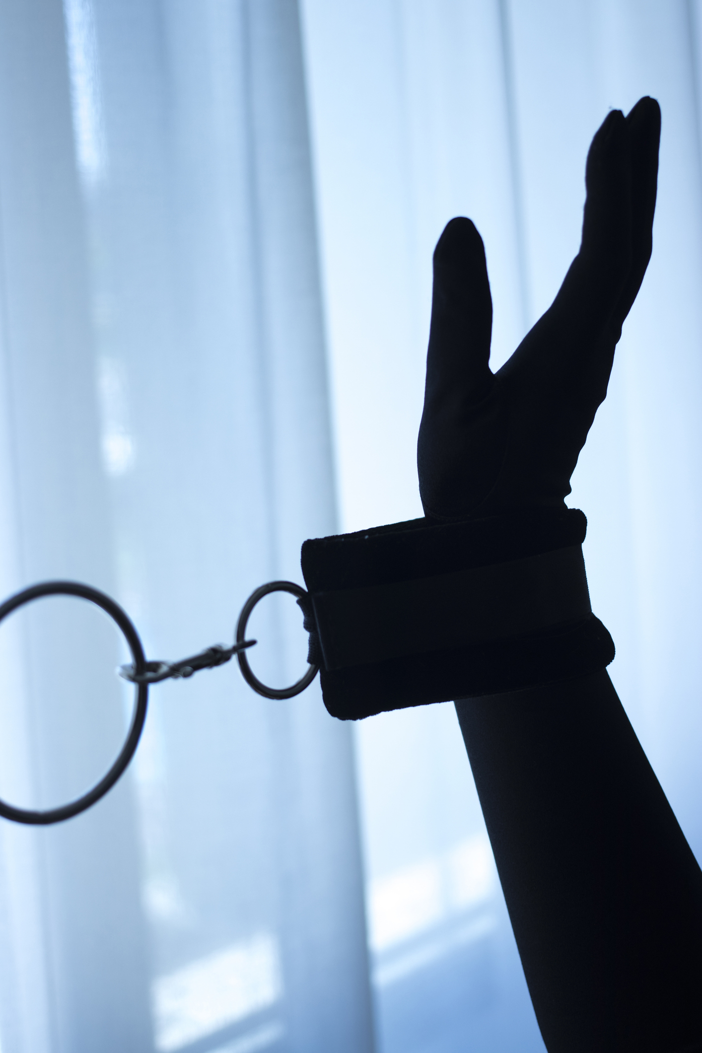 A hand in a silhouette appears to be cuffed with a restraint against a backdrop of sheer curtains