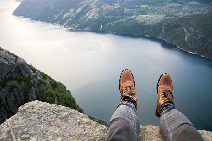 A person is sitting on the edge of a cliff overlooking a serene body of water surrounded by mountains, visible are their legs and boots