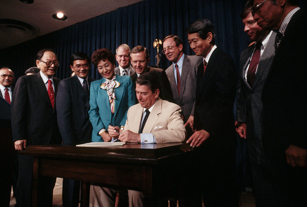 Ronald Reagan signs a document at a table surrounded by various individuals, including Norman Mineta, Robert Dole, Spark Matsunaga, and Katie Hall