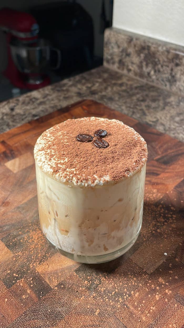 A creamy tiramisu dessert in a glass, topped with cocoa powder and coffee beans, on a wooden counter