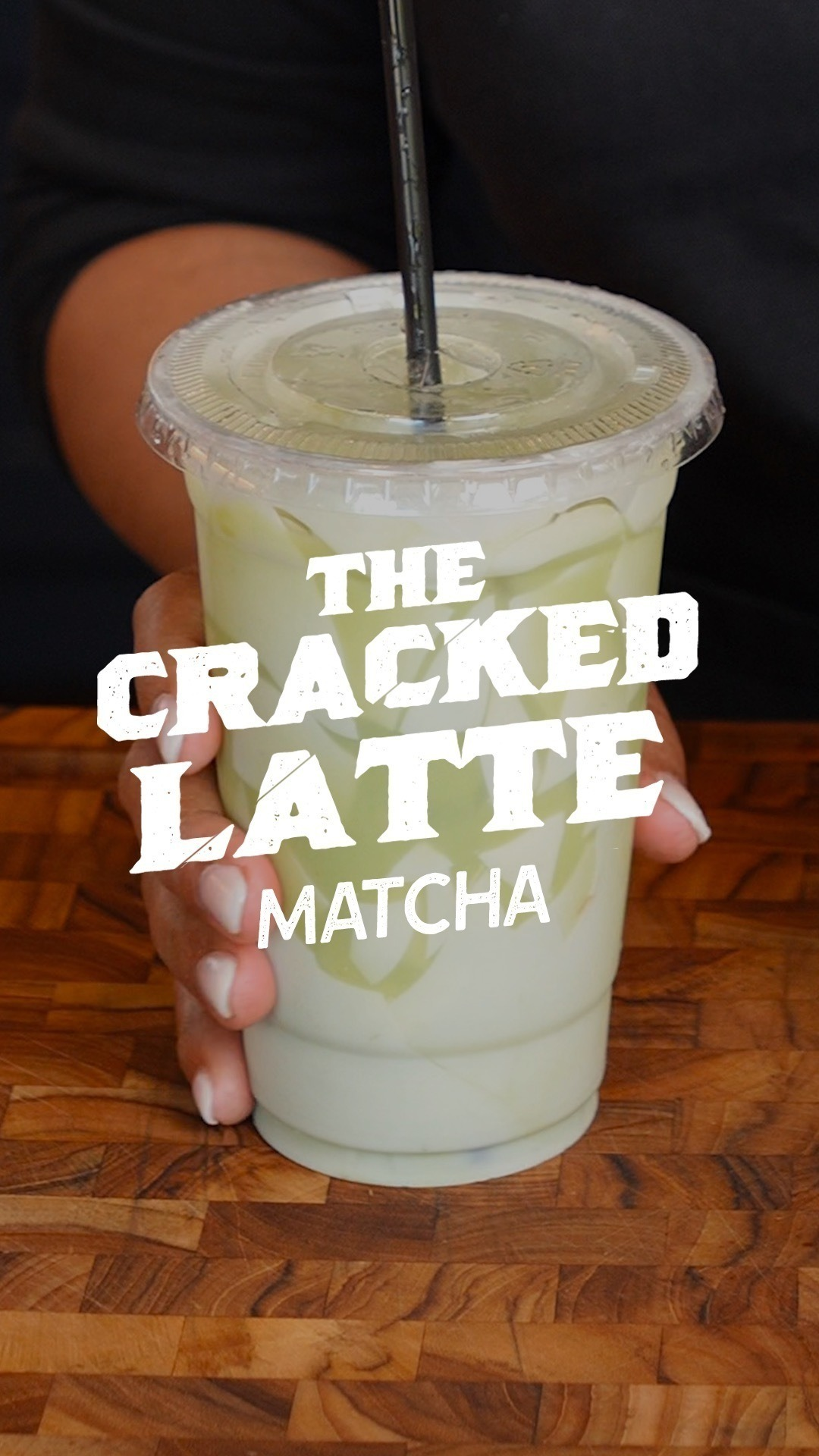A hand holds a plastic cup with a lid and straw, containing a matcha latte. Text on the image reads &quot;The Cracked Latte Matcha.&quot;