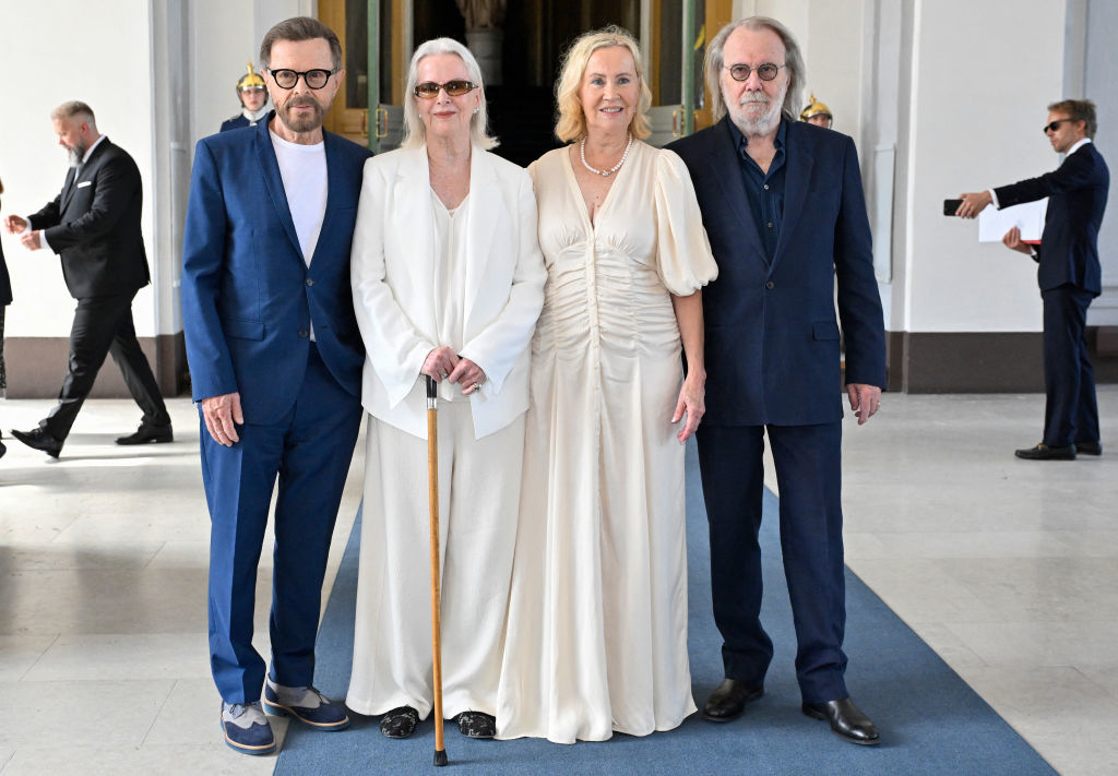 Björn Ulvaeus, Anni-Frid Lyngstad, Agnetha Fältskog, and Benny Andersson pose together indoors. Anni-Frid uses a cane, and Agnetha is in an elegant dress