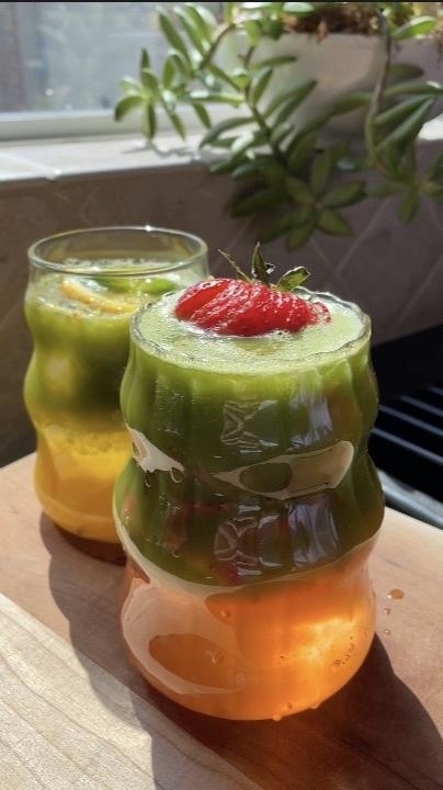 Two glasses filled with layered drinks, one topped with a strawberry, both with distinct gradient colors, placed on a wooden surface near a sunlit window with plants