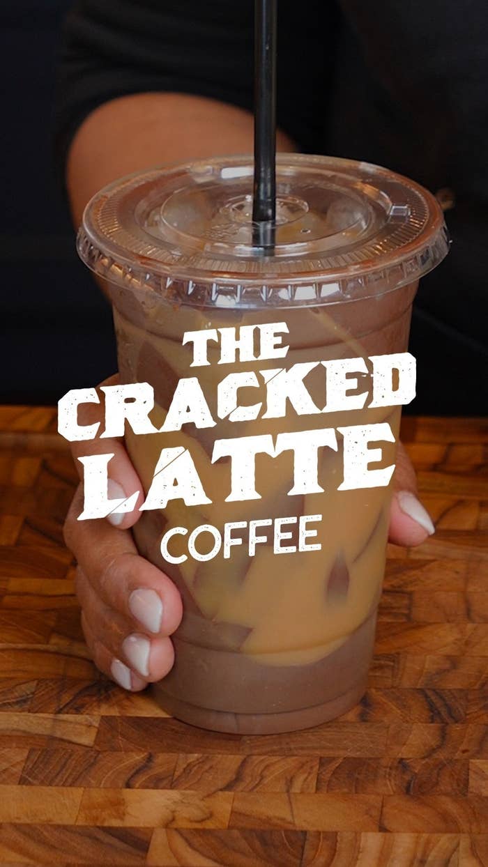 A hand holding an iced coffee cup with a straw on a wooden surface. The text on the image reads &quot;The Cracked Latte Coffee.&quot;