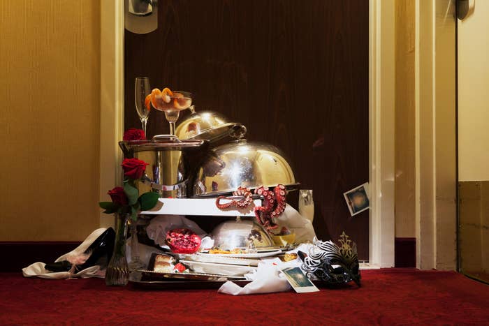 Room service trays with leftover food, champagne glasses, roses, masquerade mask, and a tiara are piled up outside a hotel room door