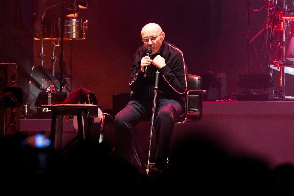 Phil Collins sitting in a chair on stage, holding a microphone mid-performance