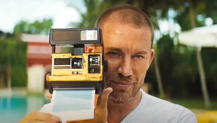 Channing Tatum holding a vintage Polaroid camera and pointing it at the viewer, set in a tropical outdoor environment in "Blink Twice"
