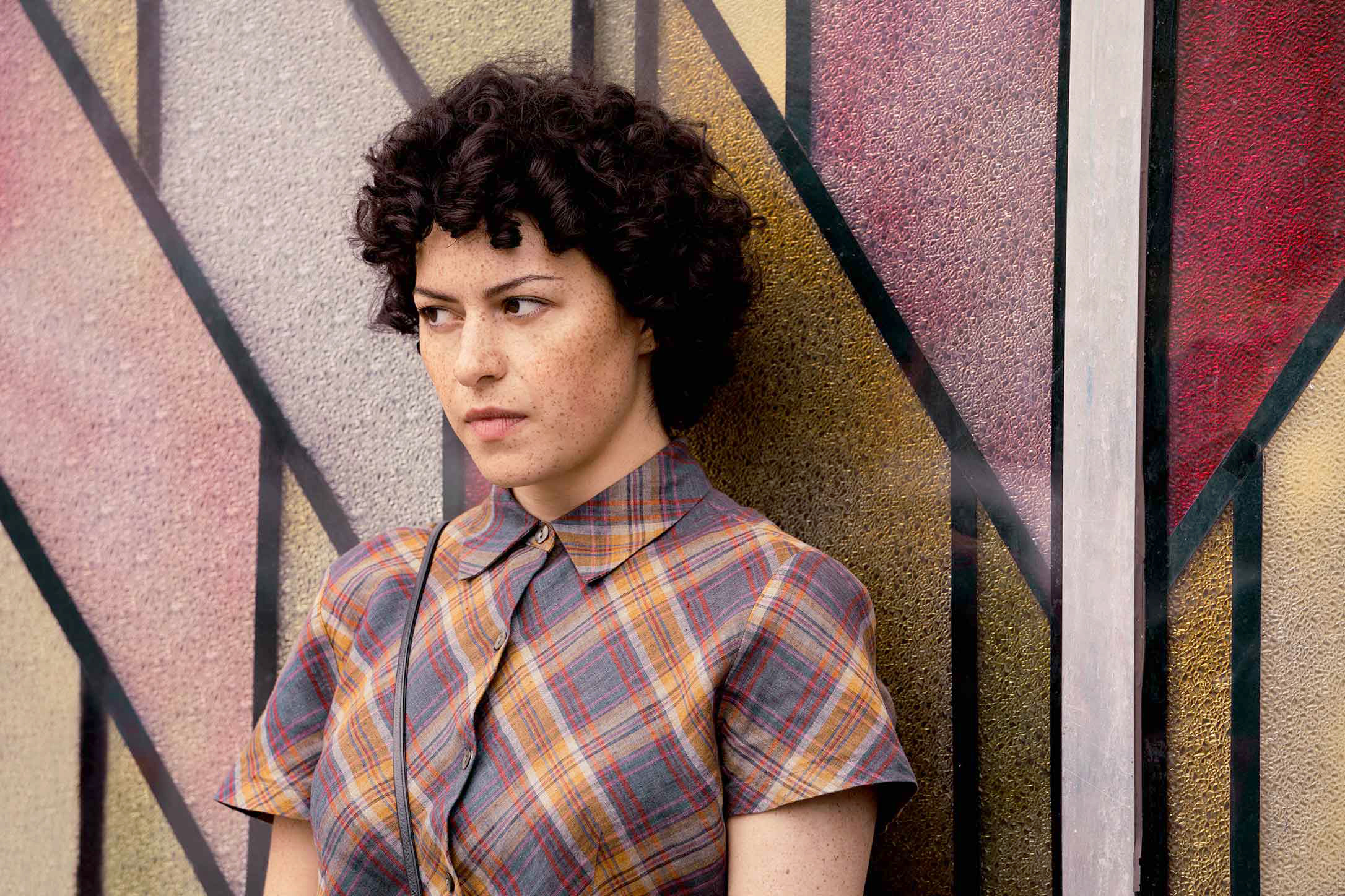 Alia Shawkat wearing a plaid shirt poses against a geometric background in "Search Party"