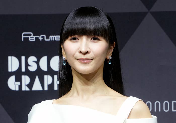 女性が右腕を前に組んで立っている。背景には"Perfume DISCOGRAPHY"と書かれたサイン。白いモダンなドレスを着用。