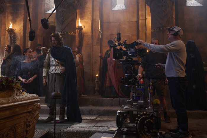 A film crew is on set, preparing to shoot a medieval scene. A man in armor and a cape stands beside a woman in a dark dress/Ema Horvath as Eärien, Lloyd Owen as Elendil