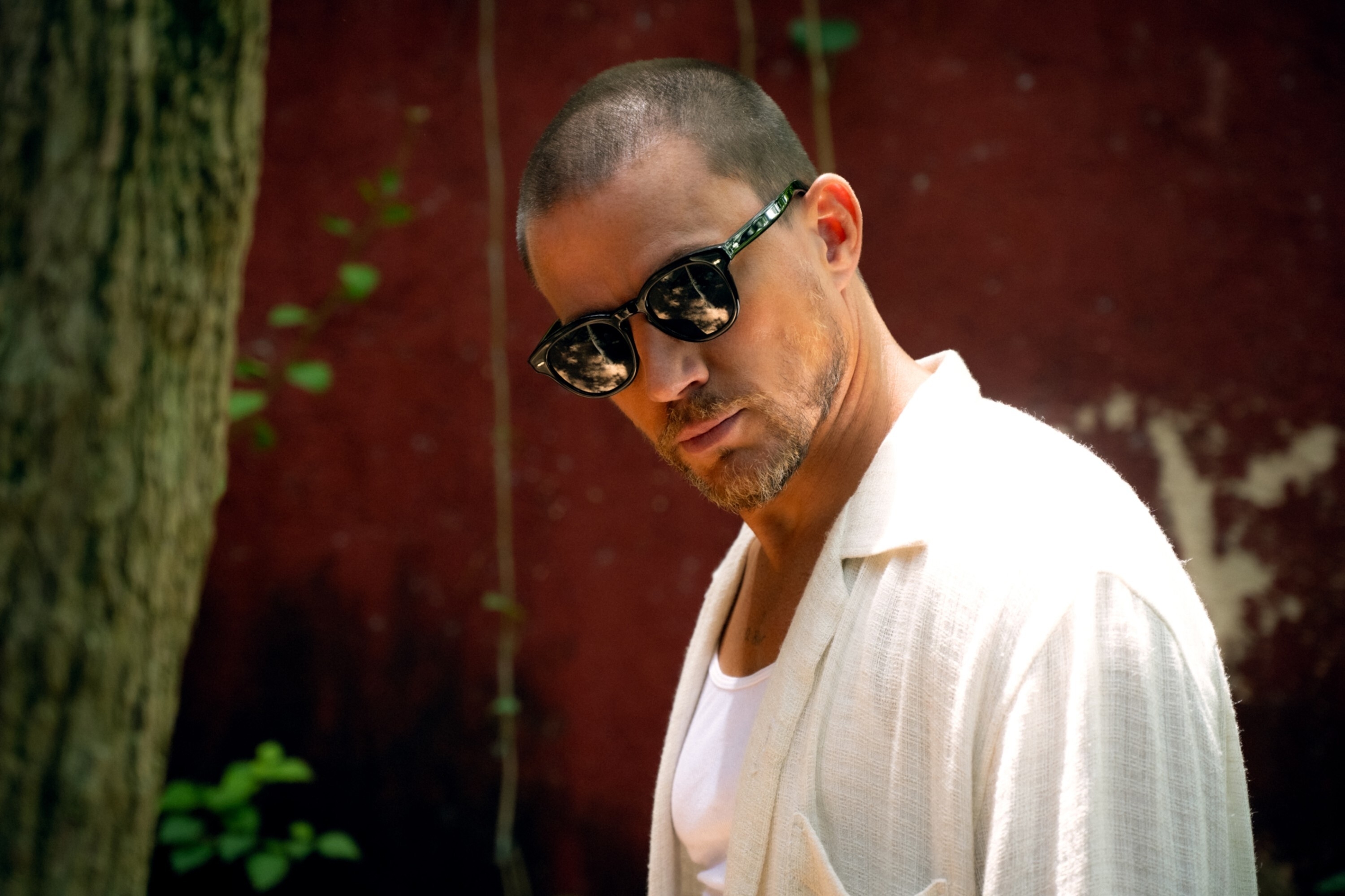 Channing Tatum wearing sunglasses and a casual, open white shirt over a white tank top, looking at the camera with a serious expression outdoors in "Blink Twice"
