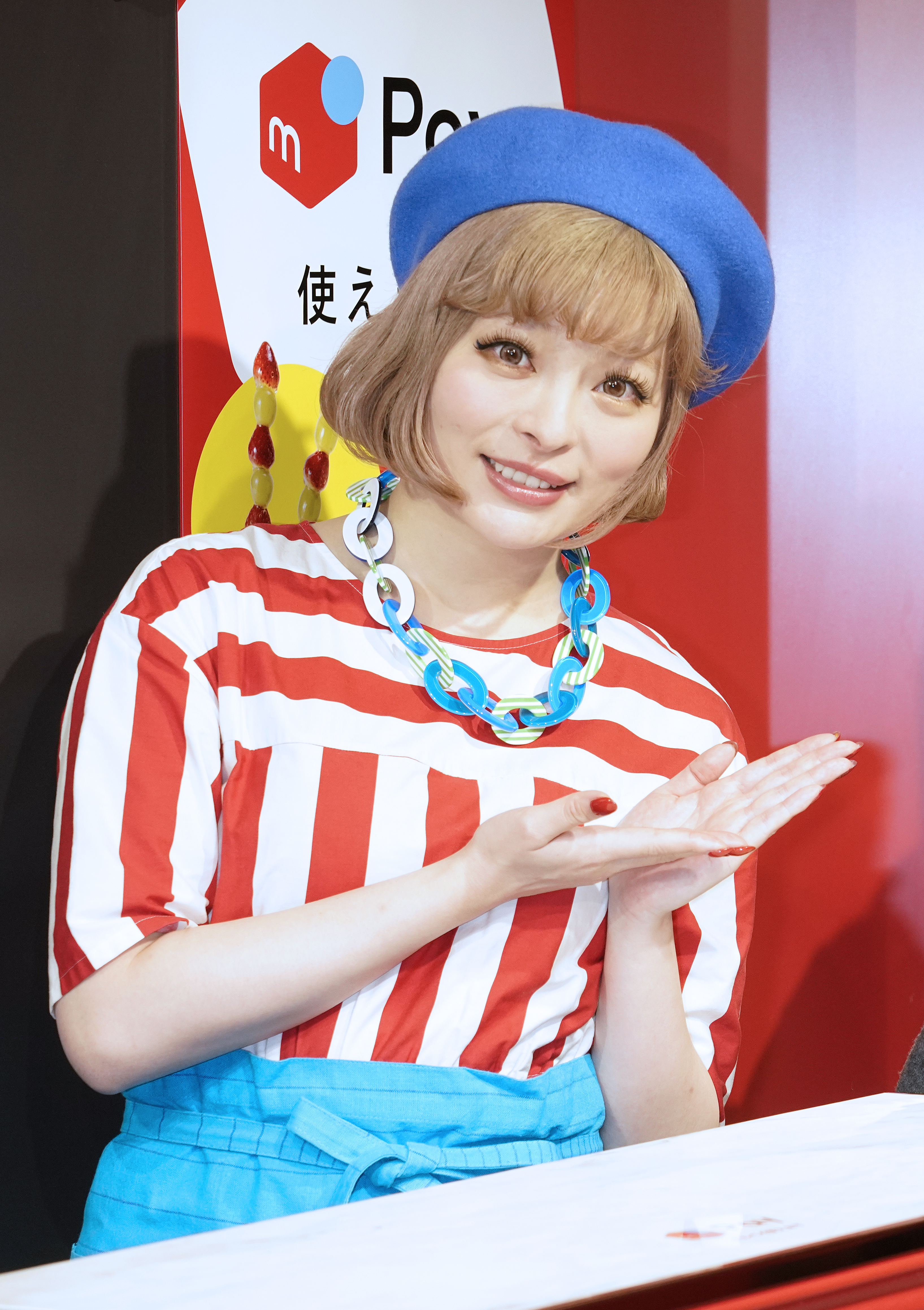 きゃりーぱみゅぱみゅが赤と白のストライプシャツ、ブルーのスカートとベレー帽を着用し、手を合わせたポーズ。背景に広告看板。