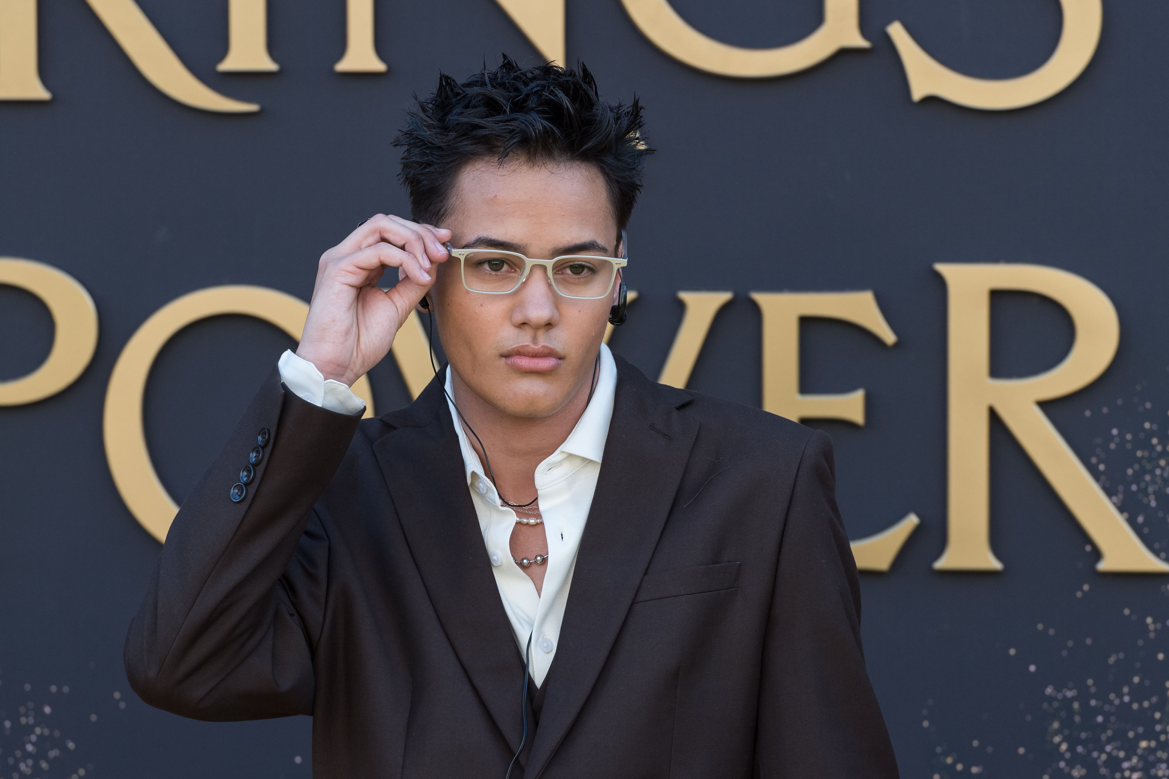 Ismael Cruz Córdova poses in a brown suit with a slightly open white shirt and fashionable glasses at a 'Rings of Power' event