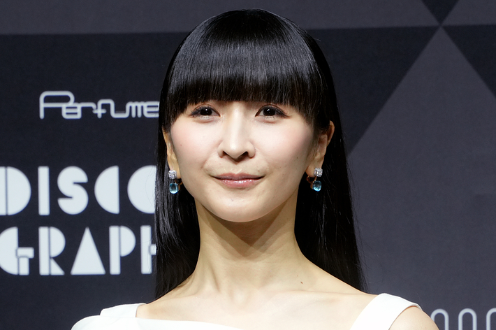 女性が右腕を前に組んで立っている。背景には&quot;Perfume DISCOGRAPHY&quot;と書かれたサイン。白いモダンなドレスを着用。