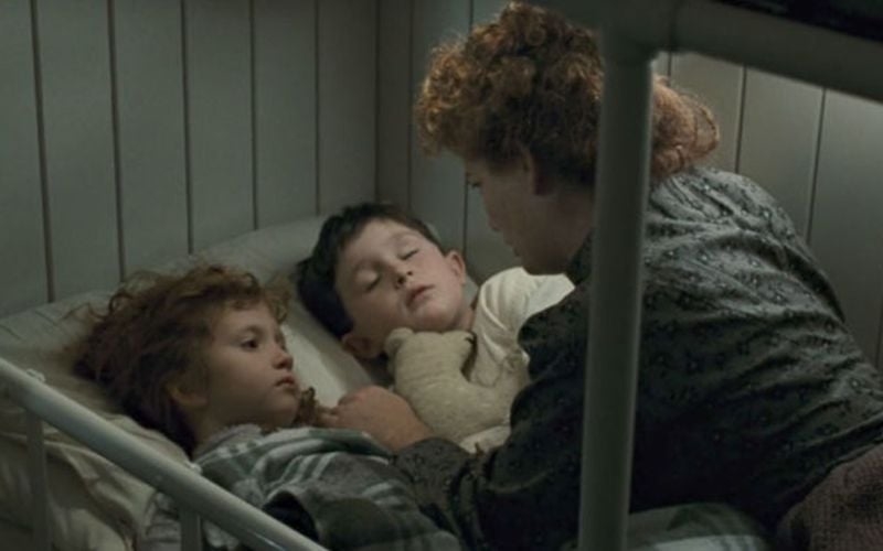 A woman with curly hair tucks two young children in bed. The children have their eyes closed, implying they are asleep. They cuddle under a blanket