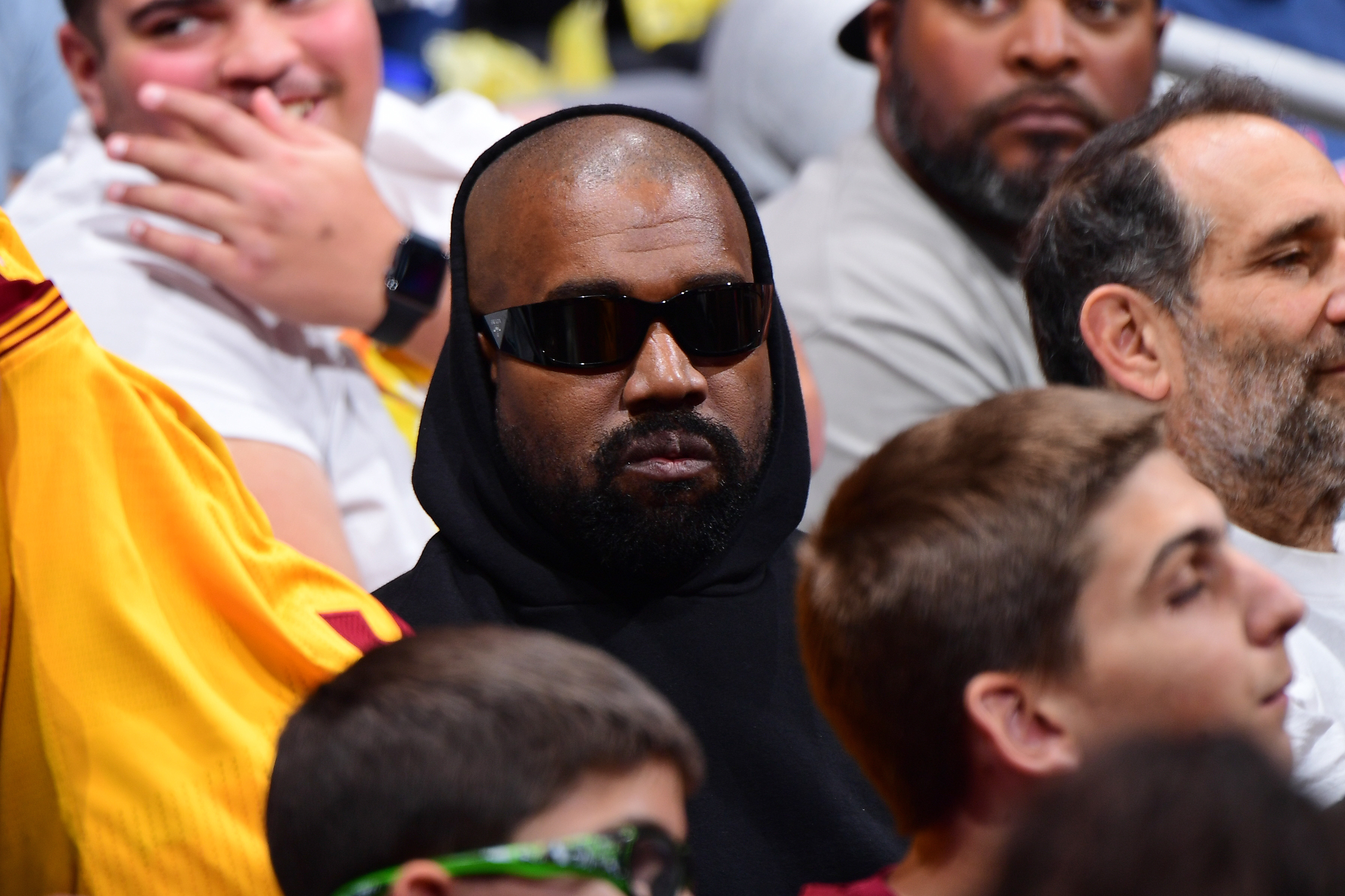 Kanye West wearing sunglasses and a hooded sweatshirt, seated among a crowd at an event