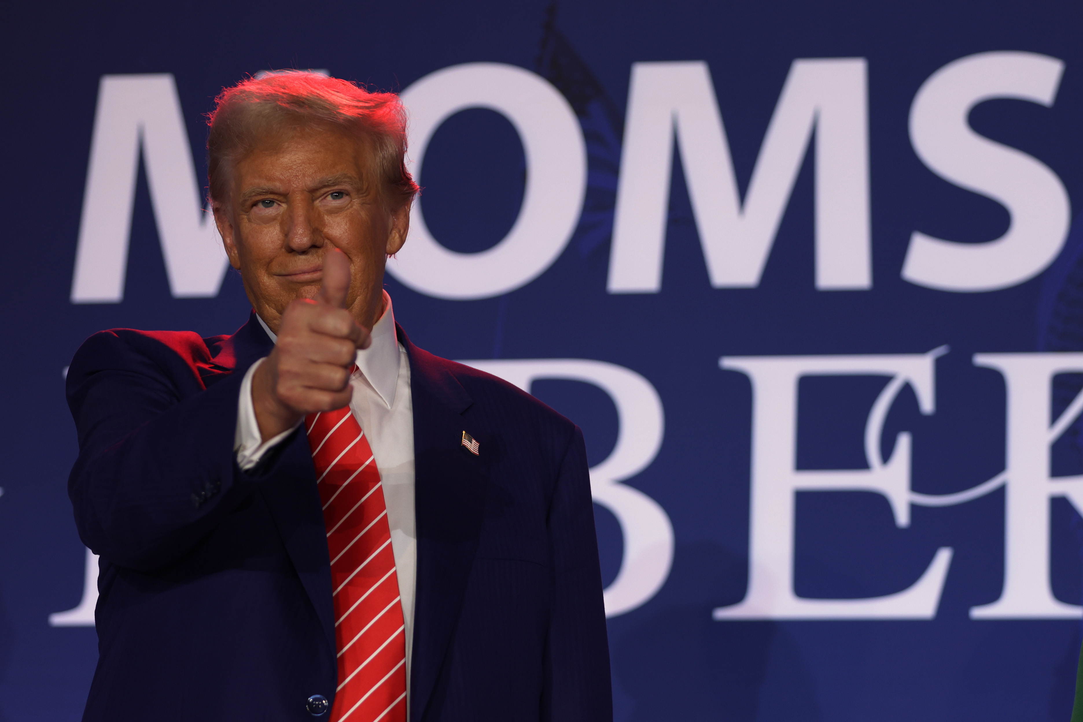 Donald Trump giving a thumbs-up gesture in front of a backdrop with the text &quot;Moms Be.&quot;