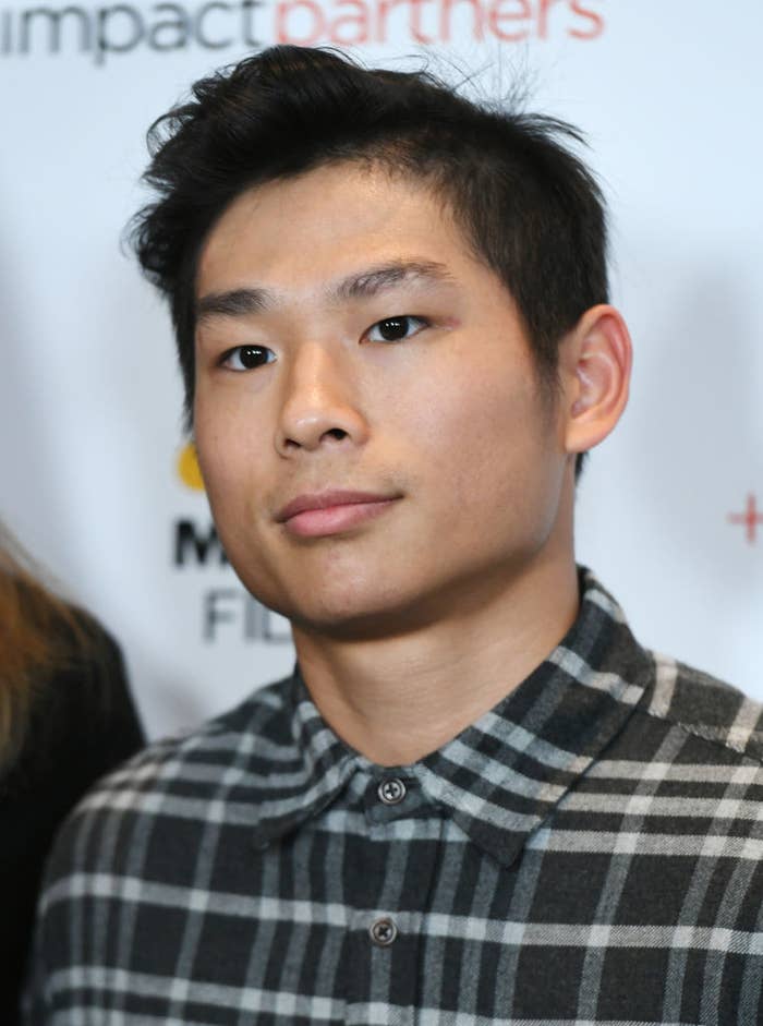 Man in a plaid shirt standing in front of a media backdrop. The backdrop has partial, unreadable text