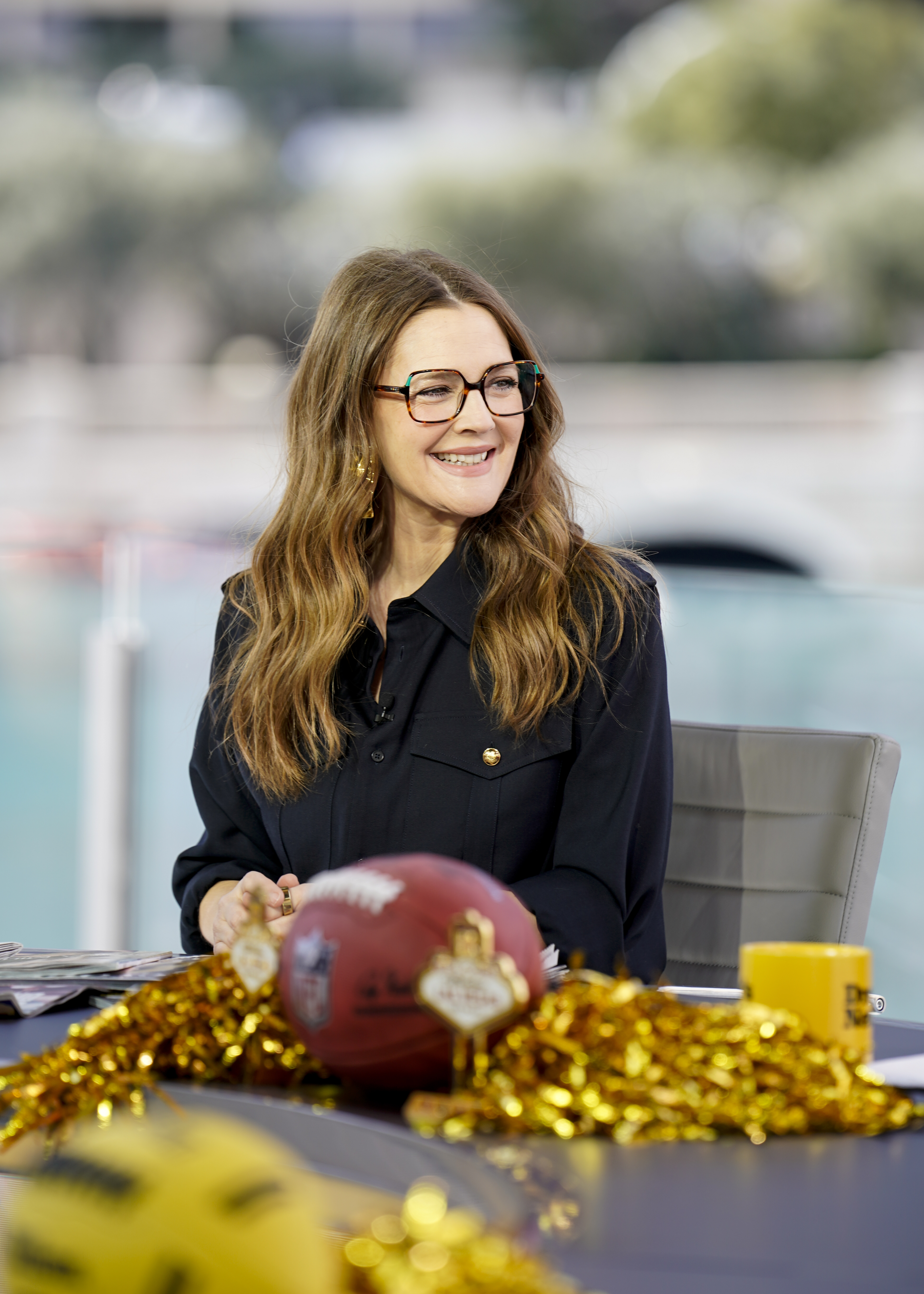 Drew Barrymore smiles at a desk with a football helmet and a football adorned with gold tinsel in front of her