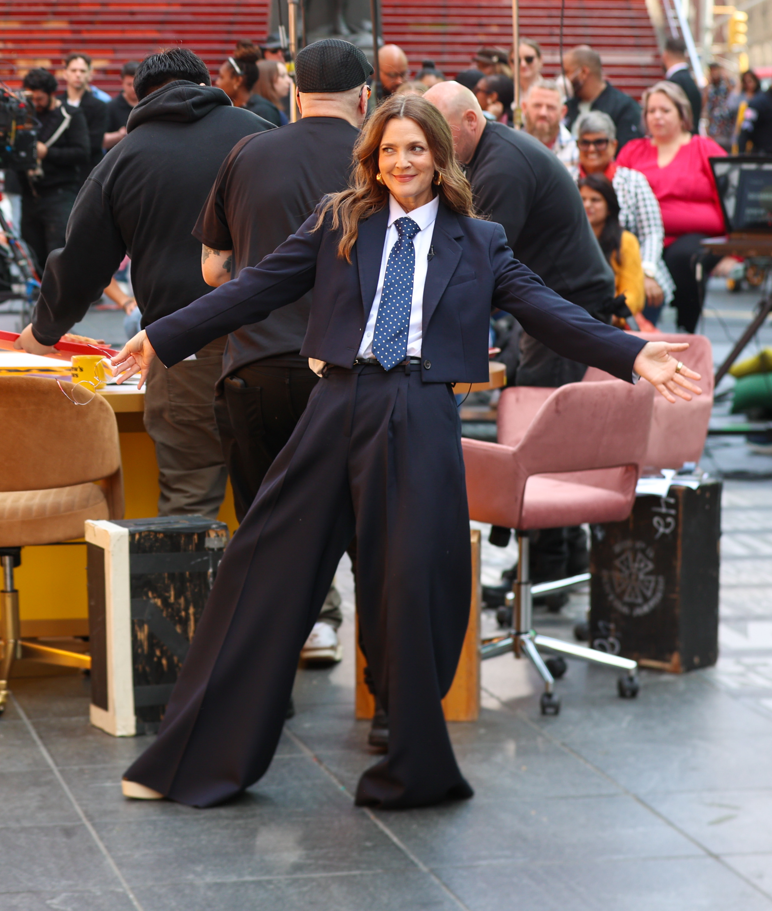 Drew Barrymore on an outdoor set wearing a baggy navy suit with a white shirt and polka dot tie, posing with arms spread wide. People are seen working around her