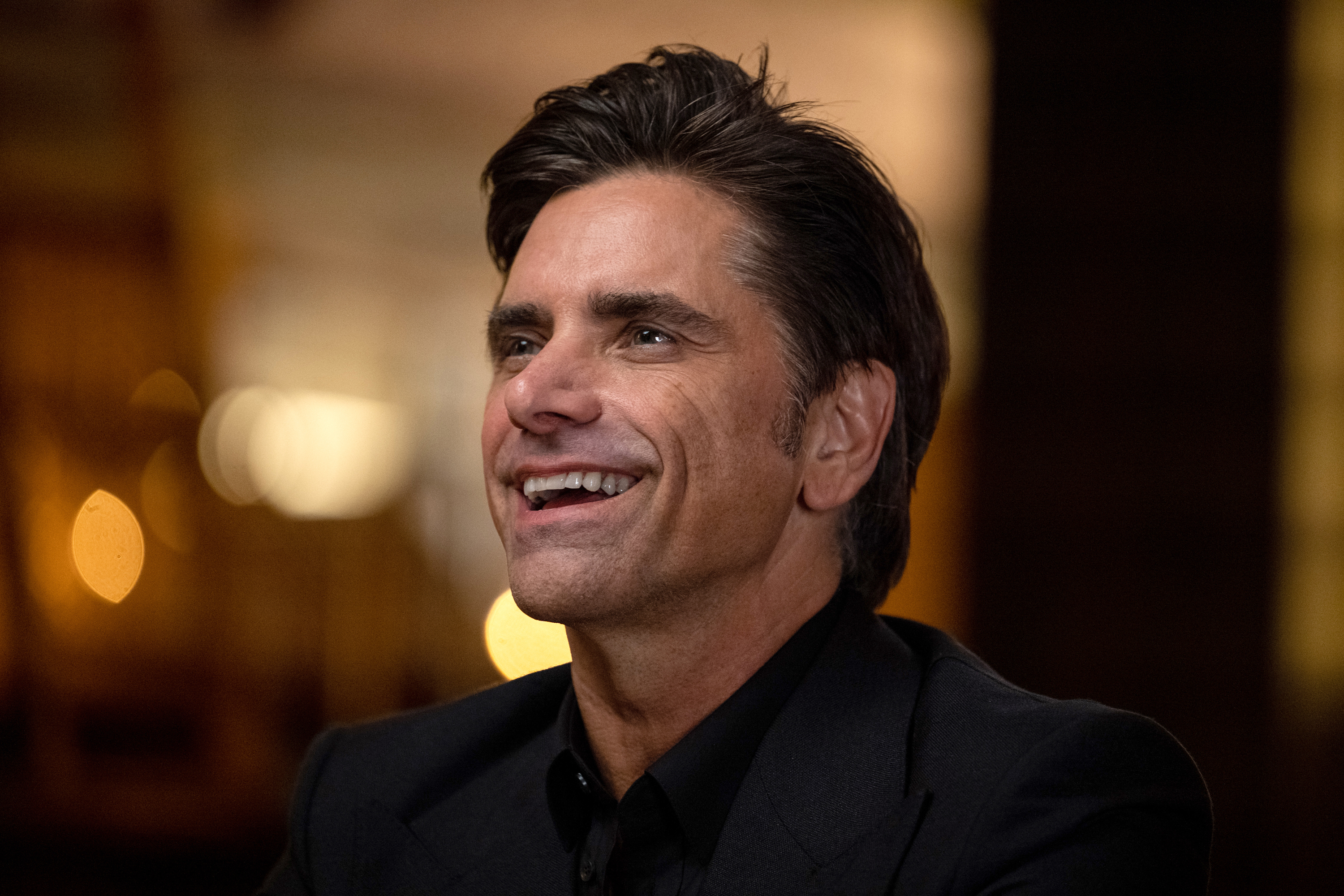 John Stamos smiling, wearing a dark suit and sitting indoors