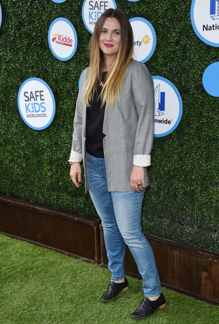 Drew Barrymore at a Safe Kids Worldwide event, wearing a black top, gray blazer, blue jeans, and black shoes