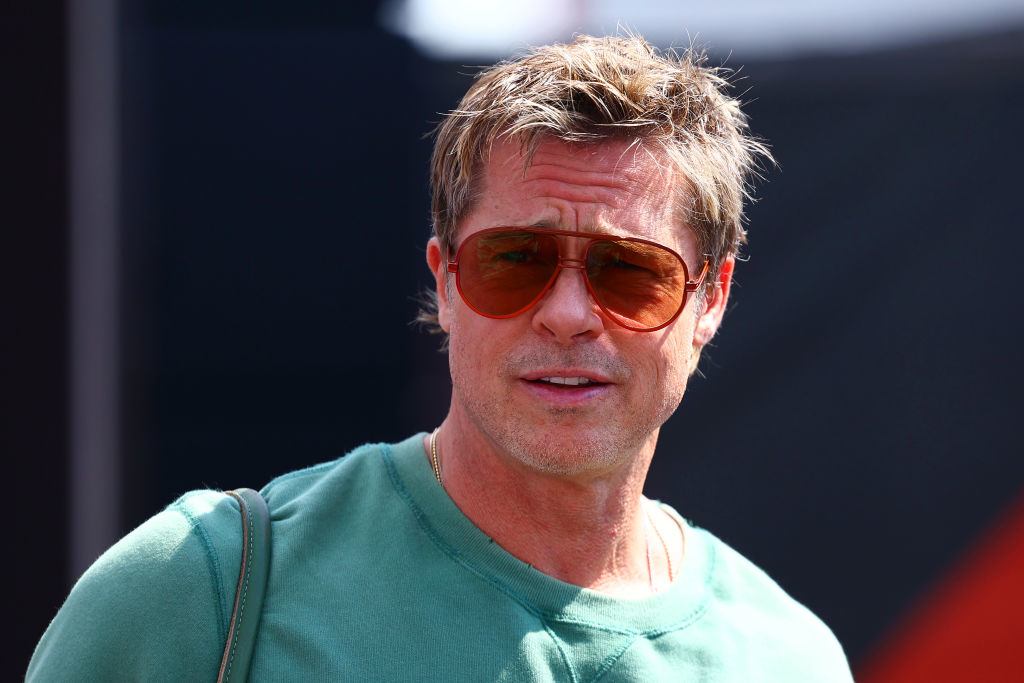Brad Pitt wears aviator sunglasses and a casual shirt, standing against a blurred background, looking slightly to the side