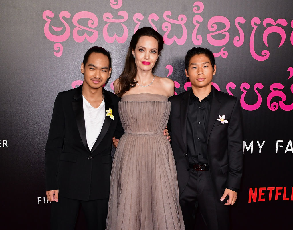Angelina Jolie stands between her sons Maddox Jolie-Pitt and Pax Jolie-Pitt at a red carpet event. All three wear formal attire with flower brooches